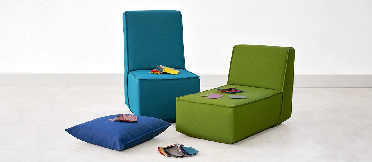 Zwei modulare Sofa-Elemente und ein Kissen mit aufgelegten Stoffmustern in einem hellen Raum, bereit zur Ansicht durch Kund:innen