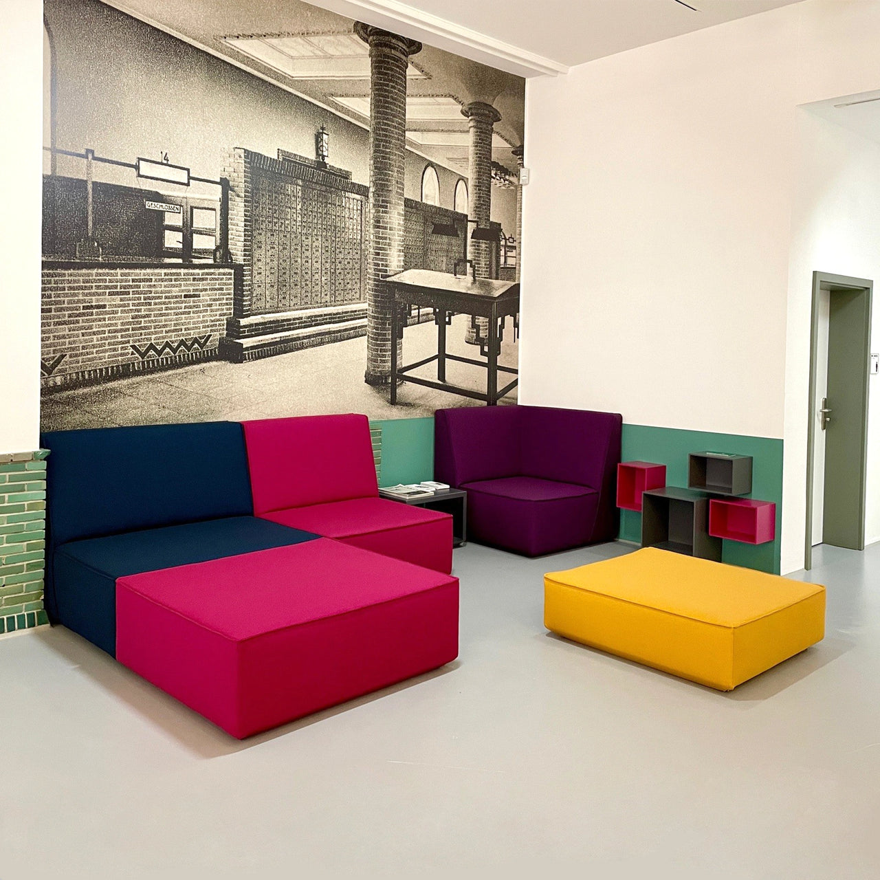 Modular seating in bold colors, complemented by small shelving modules on the wall, in front of a historical photo wall in the Mülheim an der Ruhr Art Museum.