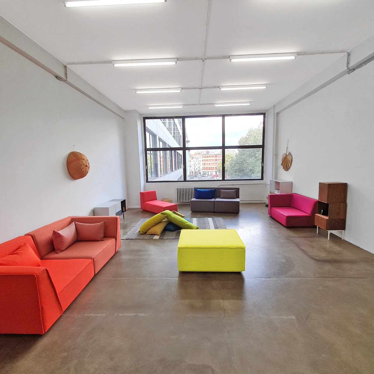 Spacious showroom in Berlin with various Cubit sofa modules in bold colors in front of a window front with a city view, complemented by sideboards and carpet on a concrete floor.