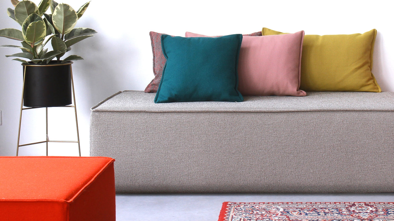 A simple sofa with a textured fabric, decorated with several differently colored cushions; in the foreground, a bright red stool, next to it a rug and a green houseplant