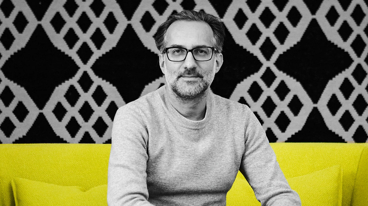 Portrait of a man with glasses and a grey sweater, sitting on a yellow Cubit Sofa in front of a graphic background