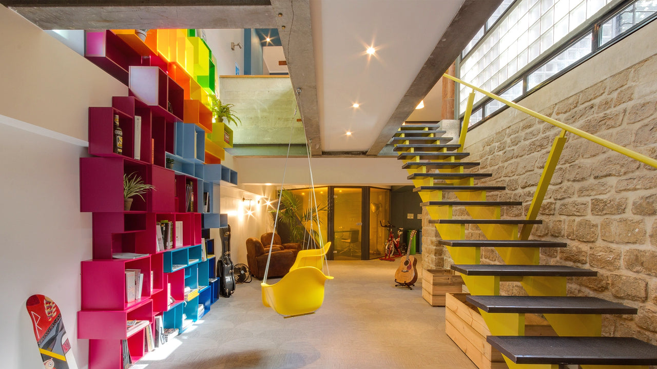 Salon lumineux et moderne avec une étagère murale aux couleurs vives composée de modules roses, bleus, jaunes et verts, une balançoire jaune et un escalier jaune avec un mur en pierre et une grande baie vitrée.