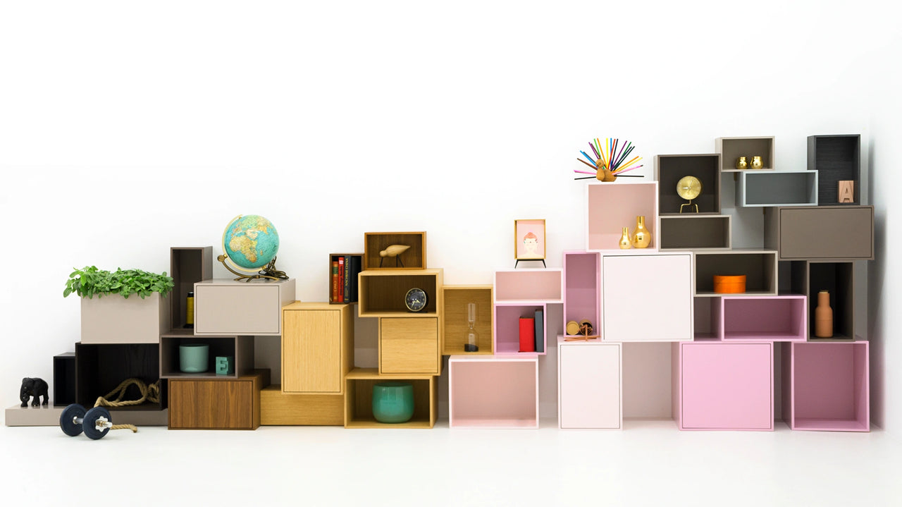 Una creativa parete di scaffali composta da scatole modulari in beige, rosa, legno e grigio, decorata con piante, libri, mappamondi, vasi dorati e oggetti di design, di fronte a una parete bianca su un pavimento bianco